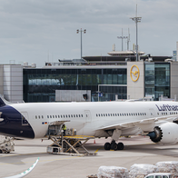 A Lufthansa aircraft parked at a jet bridge at Frankfurt Airport, with ground crew loading cargo pallets onto the tarmac.