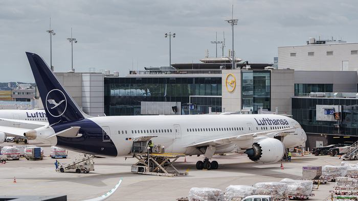 A Lufthansa aircraft parked at a jet bridge at Frankfurt Airport, with ground crew loading cargo pallets onto the tarmac.