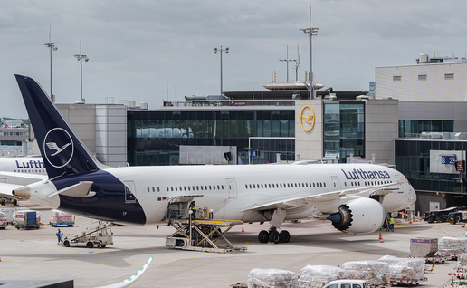 A Lufthansa aircraft parked at a jet bridge at Frankfurt Airport, with ground crew loading cargo pallets onto the tarmac.