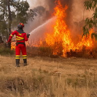 UME firefighter in red uniform sprays water on intense wildfire flames in a dry grassy field.