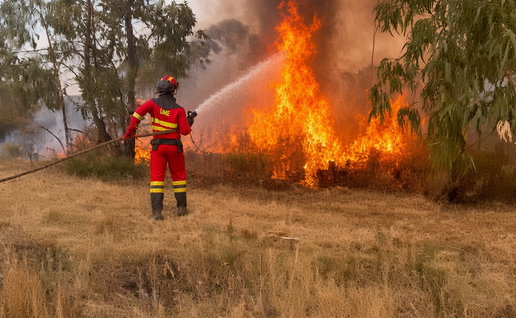 UME firefighter in red uniform sprays water on intense wildfire flames in a dry grassy field.