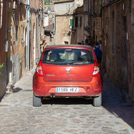 A red car driving through a narrow, flower-lined cobbled street between stone village houses.