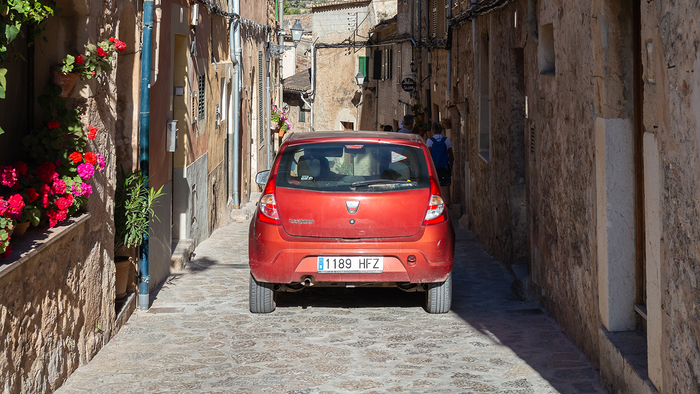 A red car driving through a narrow, flower-lined cobbled street between stone village houses.