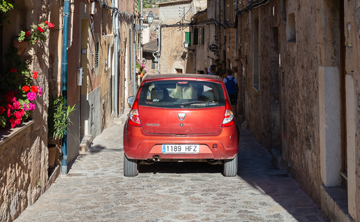 A red car driving through a narrow, flower-lined cobbled street between stone village houses.