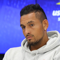 Nick Kyrgios in a grey hoodie looking down thoughtfully at a tennis press conference with a blue backdrop.