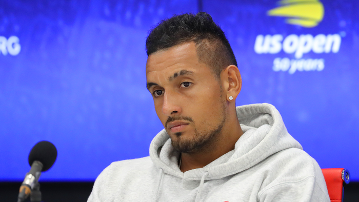 Nick Kyrgios in a grey hoodie looking down thoughtfully at a tennis press conference with a blue backdrop.