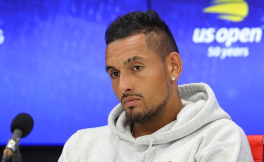 Nick Kyrgios in a grey hoodie looking down thoughtfully at a tennis press conference with a blue backdrop.