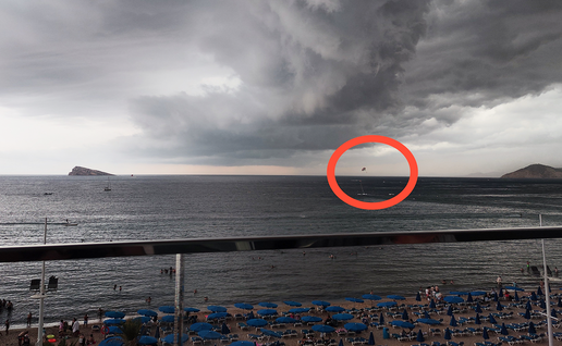 A parasail canopy circled in red above the sea off Benidorm's Levante beach, with heavy storm cloud overhead and sunbeds along the sand below.
