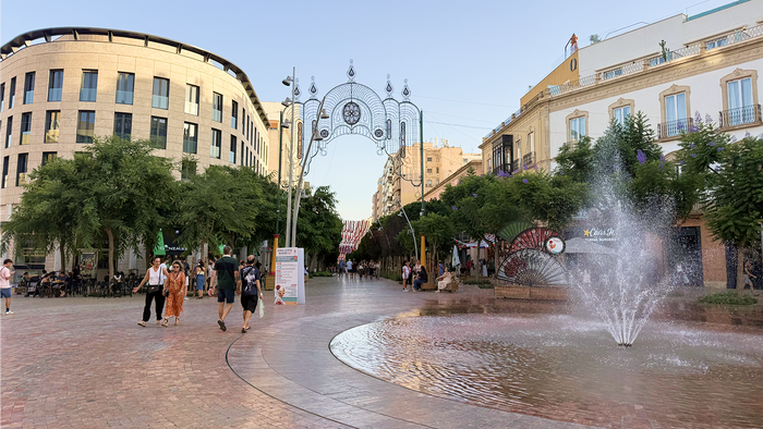 Pedestrians walking along Almería's pedestrianised Paseo, with the Feria's illuminated entrance arch and a fountain at Puerta de Purchena.