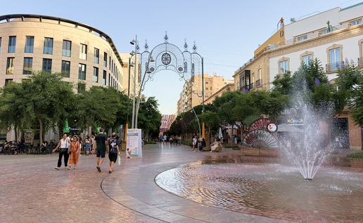 Pedestrians walking along Almería's pedestrianised Paseo, with the Feria's illuminated entrance arch and a fountain at Puerta de Purchena.