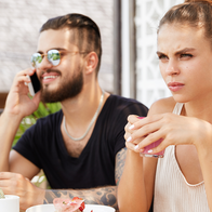 A man looks at his phone while a woman sits with her arms crossed at an outdoor café table with coffee and croissants.