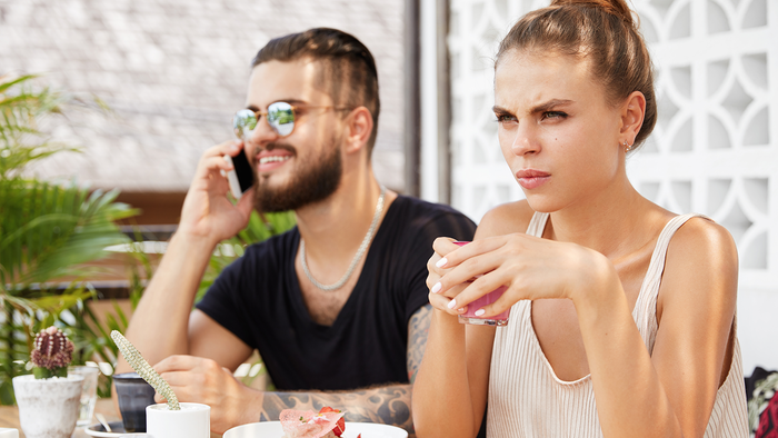 A man looks at his phone while a woman sits with her arms crossed at an outdoor café table with coffee and croissants.