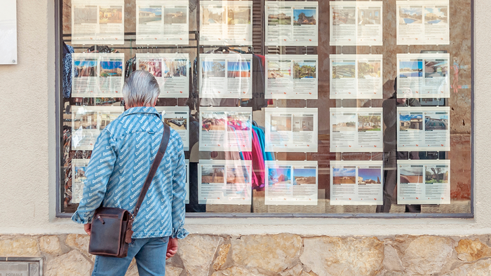 Person looking at rows of property listings displayed in a Spanish estate-agent window.