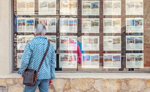 Person looking at rows of property listings displayed in a Spanish estate-agent window.