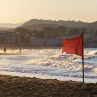A red warning flag flutters on a beach at sunset, with waves breaking in the background and people walking along the shore.
