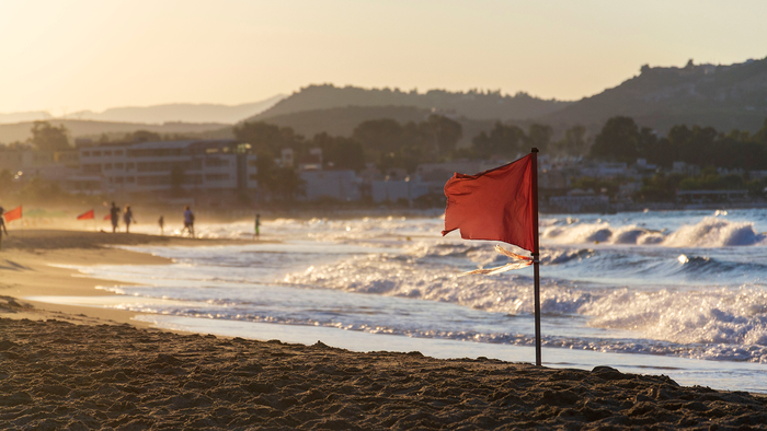 A red warning flag flutters on a beach at sunset, with waves breaking in the background and people walking along the shore.