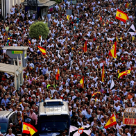 Large crowd of people waving Spanish flags at a street demonstration, with police vans in the foreground.