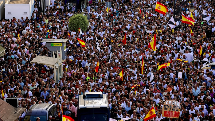 Large crowd of people waving Spanish flags at a street demonstration, with police vans in the foreground.