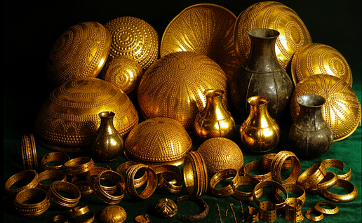 Collection of ancient gold bowls, vases, bracelets, and ornaments arranged on a dark green surface.