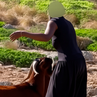 Woman in dark dress with face censored, standing beside a brown goat on rocky terrain.