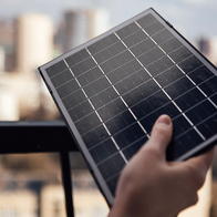 A hand holding a small solar panel up to a balcony railing, with a city skyline in the background.