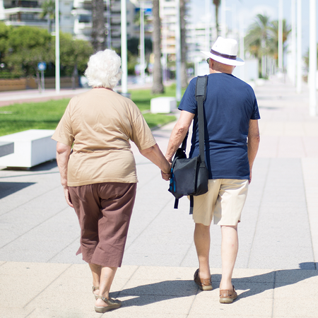 Elderly couple holding hands as they walk down a Spanish seafront.