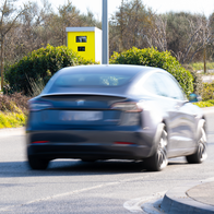 A dark car blurred with motion drives away from the camera past a yellow fixed speed camera box on a pole, on a road bordered by bushes and trees.