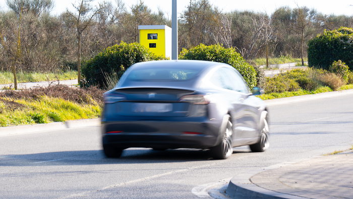 A dark car blurred with motion drives away from the camera past a yellow fixed speed camera box on a pole, on a road bordered by bushes and trees.