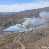 Aerial photo of thick white and grey smoke rising from a wildfire in hilly terrain near wind turbines at Las Tres Villas, Almería.