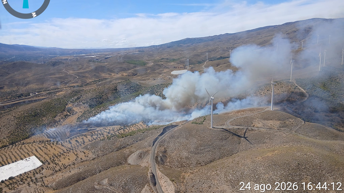 Aerial photo of thick white and grey smoke rising from a wildfire in hilly terrain near wind turbines at Las Tres Villas, Almería.