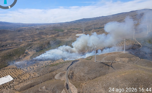 Aerial photo of thick white and grey smoke rising from a wildfire in hilly terrain near wind turbines at Las Tres Villas, Almería.