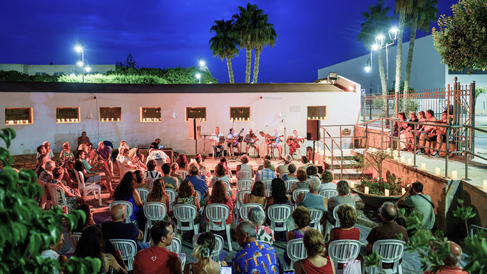 Audience gathers between candlelit walls in front of a stage of musicians performing at Vera's Plaza de los Cuatro Caños.