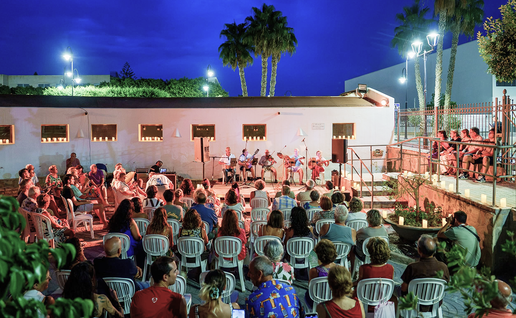 Audience gathers between candlelit walls in front of a stage of musicians performing at Vera's Plaza de los Cuatro Caños.
