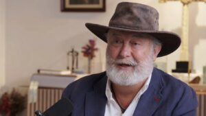 A man in a blue coat and white shirt with a white beard and moustache wearing a hat 