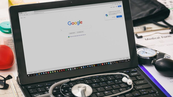 A stethoscope resting on a laptop keyboard displaying the Google search homepage surrounded by medical equipment, illustrating online symptom searching.