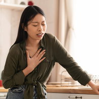 A young woman standing in her kitchen holding her hand to her chest with a pained expression, illustrating symptoms of severe heartburn or acid reflux.