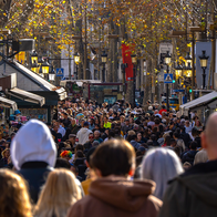 A dense crowd of pedestrians walking along a tree-lined pedestrian street in Barcelona, lined with kiosks and lamp posts.