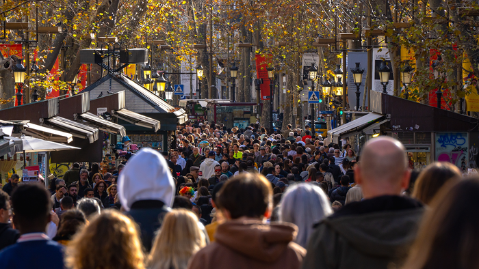 A dense crowd of pedestrians walking along a tree-lined pedestrian street in Barcelona, lined with kiosks and lamp posts.
