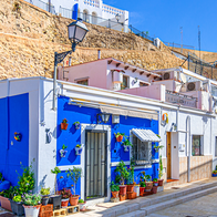 A narrow street of blue and white houses with potted plants, beneath a hillside stone wall and a whitewashed lookout point.