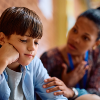 A young boy looks downcast while an adult crouches beside him, offering comfort in a school corridor.