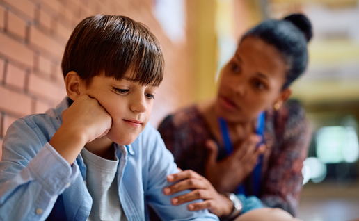 A young boy looks downcast while an adult crouches beside him, offering comfort in a school corridor.