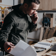 A self-employed tradesman on the phone while reviewing paperwork, an invoice and a calculator at his workshop workbench.