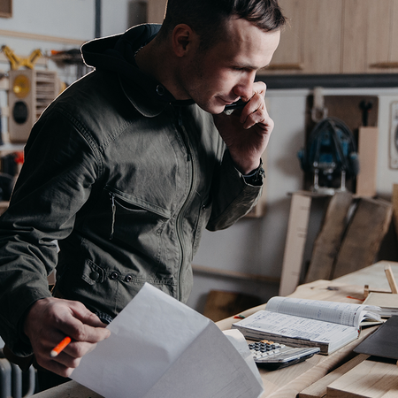 A self-employed tradesman on the phone while reviewing paperwork, an invoice and a calculator at his workshop workbench.