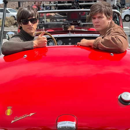 Two young actors in 1960s-style clothing sit in a red vintage Austin Healey Sprite convertible on a film set in Tenerife, with camera equipment and crew visible around them.