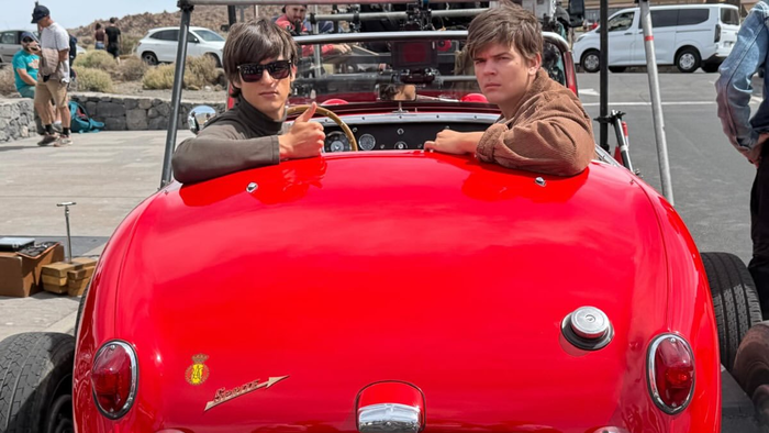 Two young actors in 1960s-style clothing sit in a red vintage Austin Healey Sprite convertible on a film set in Tenerife, with camera equipment and crew visible around them.