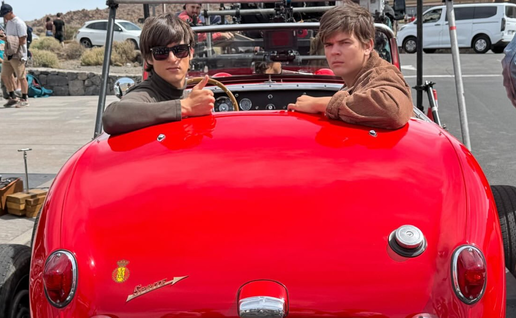 Two young actors in 1960s-style clothing sit in a red vintage Austin Healey Sprite convertible on a film set in Tenerife, with camera equipment and crew visible around them.
