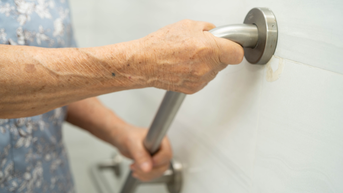 An elderly person's hand gripping a stainless steel grab rail fitted to a bathroom wall, with a mobility frame nearby.