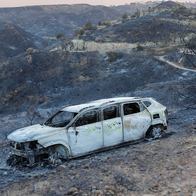 A burnt-out car sits abandoned on a fire-blackened hillside under a smoky sky.