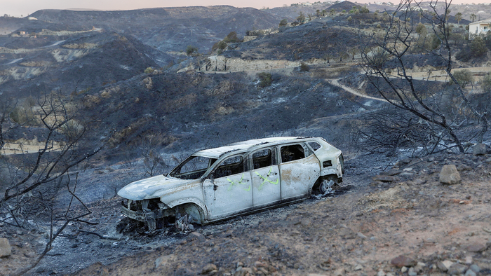 A burnt-out car sits abandoned on a fire-blackened hillside under a smoky sky.