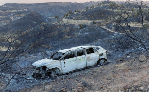 A burnt-out car sits abandoned on a fire-blackened hillside under a smoky sky.